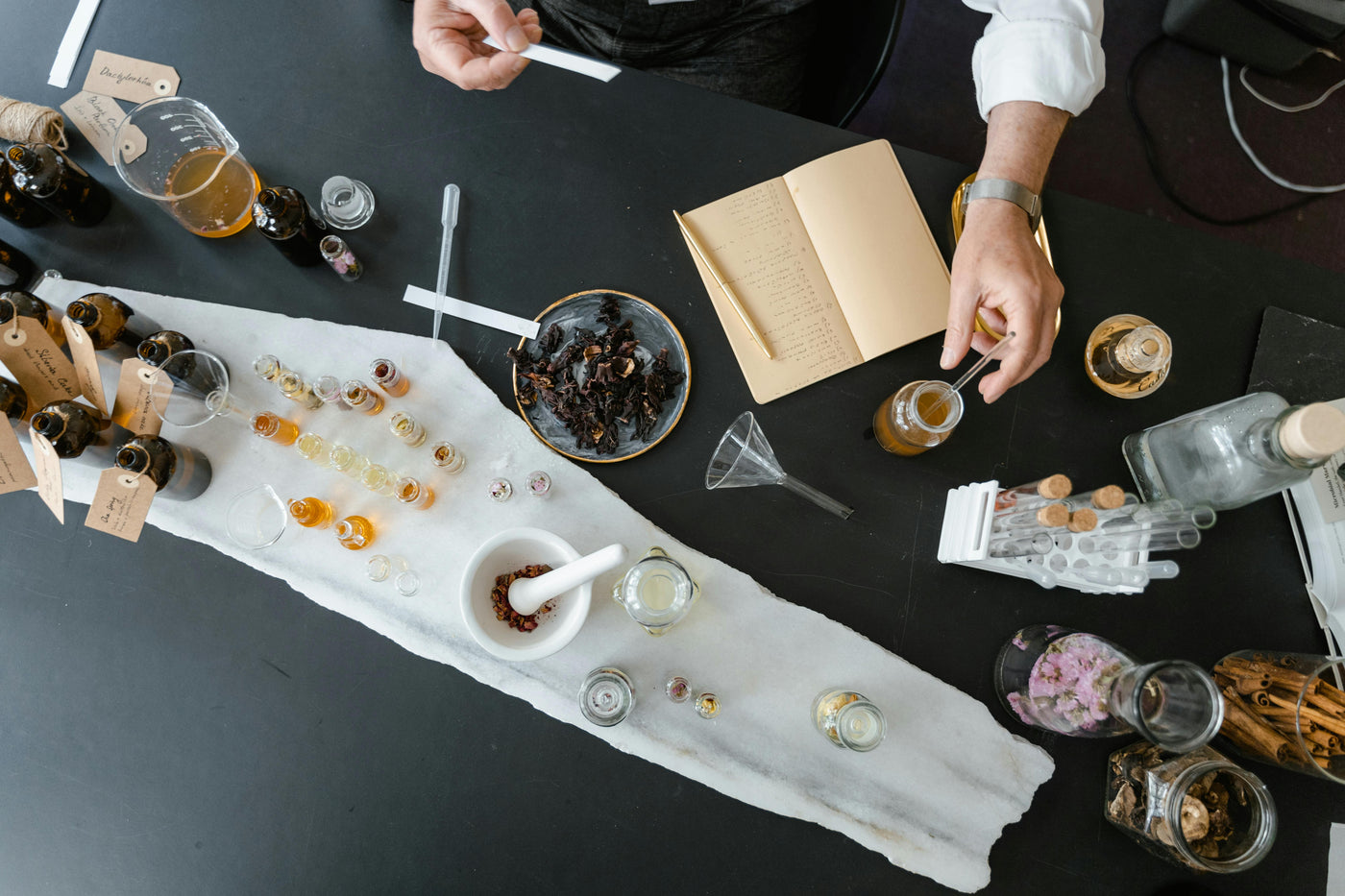 Top view of a formulator blending natural ingredients and essential oils in a lab setting, with dried botanicals, glass vials, test tubes, and handwritten notes on a dark table