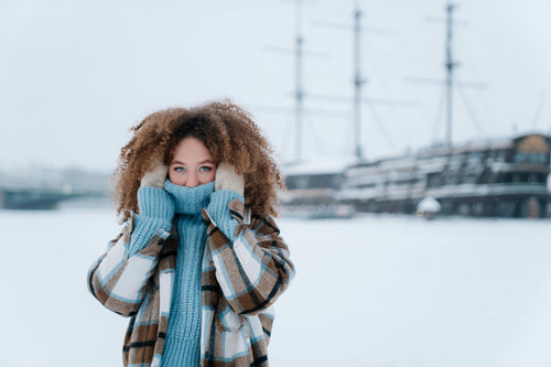 Young woman with curly hair bundled up in a plaid coat and blue knit sweater, standing outside in snowy weather with a ship in the background.